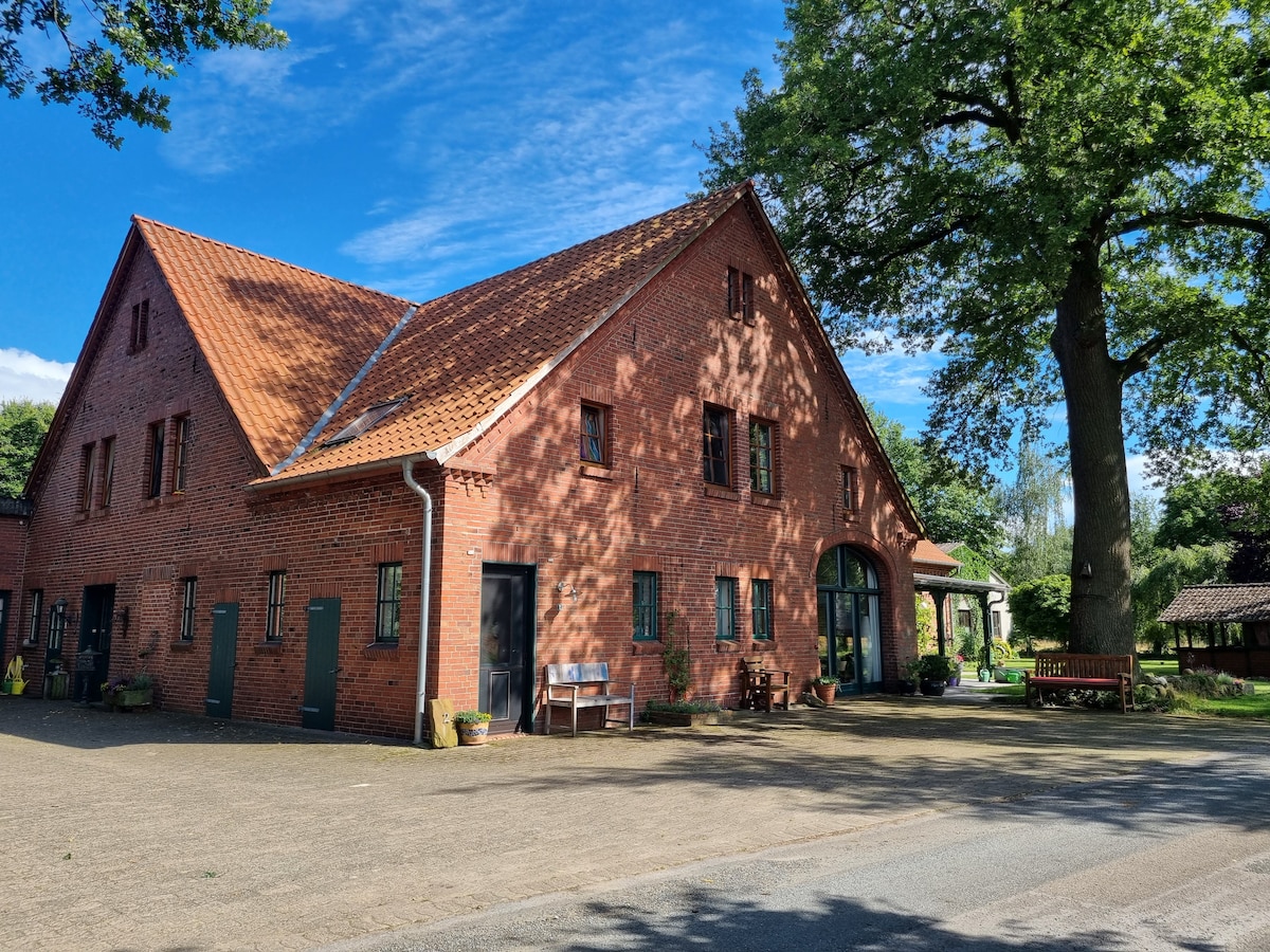 A charming red brick building with a sloping roof is featured in this image, surrounded by lush greenery. Large windows and a welcoming entrance are visible, while benches provide seating along the pathway. A tall tree offers shade nearby, enhancing the serene outdoor environment.