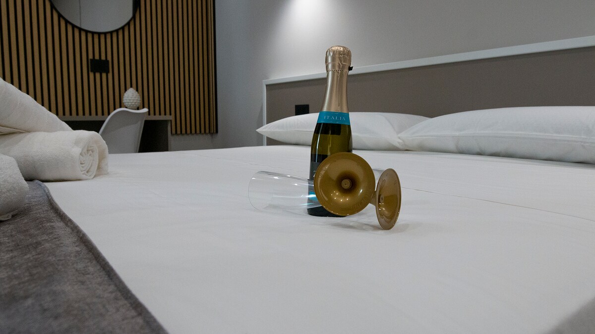 A spacious bed is centered in the room, featuring a neutral-colored bedspread. A neatly placed bottle of sparkling wine stands on the bed alongside two elegant glasses. Soft, white towels are visible nearby, contributing to the room's minimalist design.