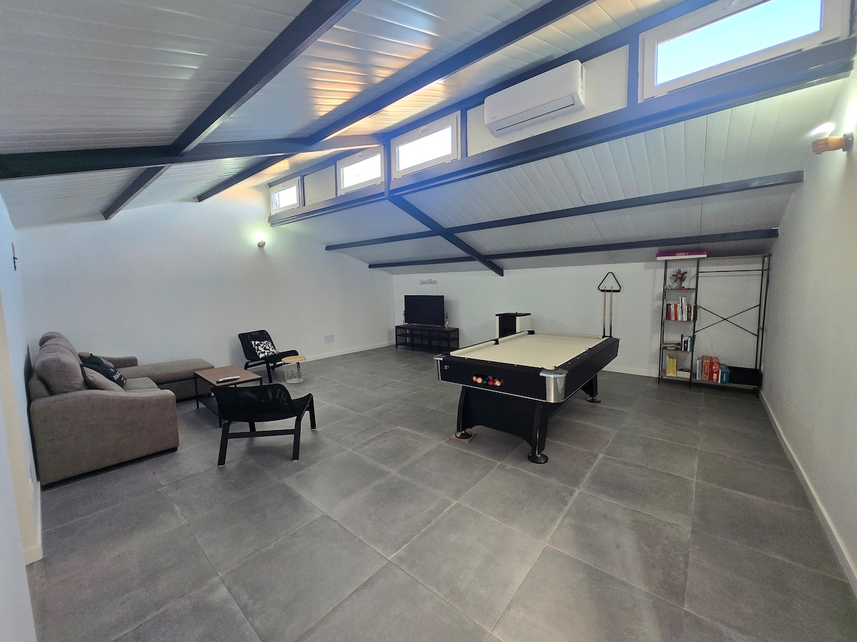An expansive recreational area features a professional billiard table centrally located. Soft seating is arranged near a wall-mounted Smart TV, with natural light filtering in through multiple windows. Air conditioning and functional shelving are also present, providing comfort and utility.