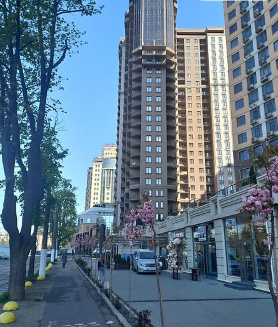 A modern residential complex is featured, showcasing a tall building with a mix of textures and materials. Trees line the walkway, which is bordered by vibrant pink flowering plants. On the ground level, shops and businesses are visible, providing accessible amenities for residents.