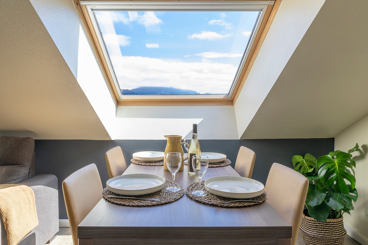 A dining area is featured, with a wooden table set for four. Plates and glasses are arranged on placemats. A large skylight provides natural light, illuminating the space and offering views of the sky. A decorative plant adds a touch of greenery.