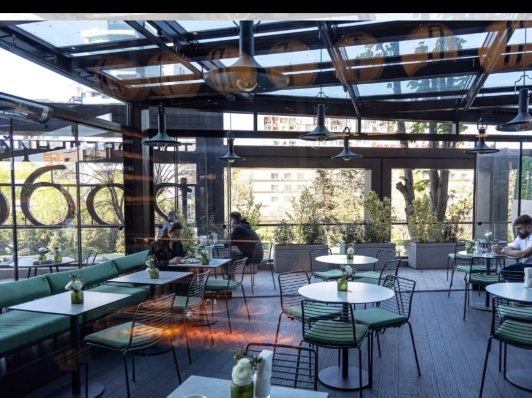 An indoor-outdoor dining area features several round tables accompanied by green chairs. Large glass panels allow natural light to fill the space. Potted plants line the perimeter, enhancing the vibrant atmosphere. Guests are seated, enjoying their meals amidst the greenery and open views.