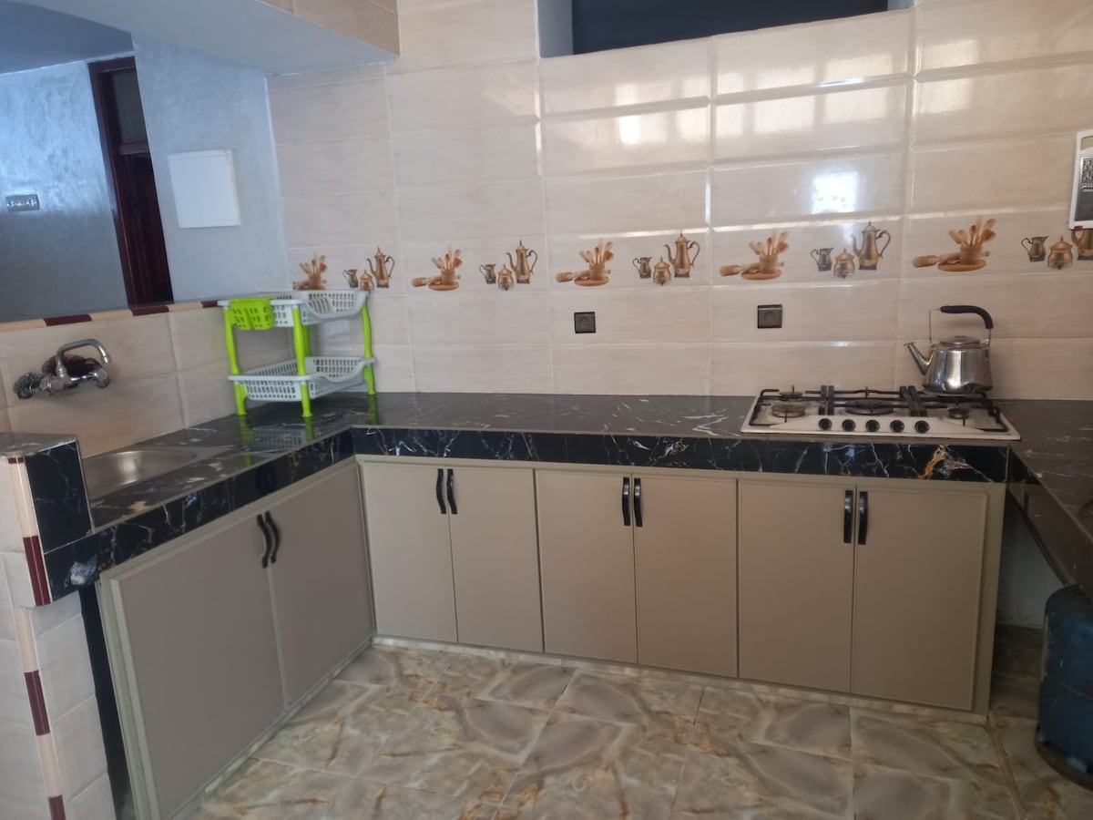 A functional kitchen area features sleek, dark marble countertops complemented by light beige cabinetry. A stovetop and kettle are present on the surface, while a small green rack provides storage. Decorative tiles with a nature theme line the walls, adding a subtle touch.