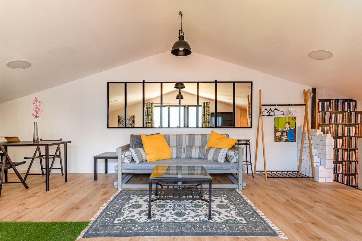 The spacious living area features a light-colored sofa with yellow cushions, positioned facing a coffee table on a patterned rug. A large mirror reflects natural light, while a table and chairs are arranged for dining. Decorative elements include art and bookshelves on the walls.
