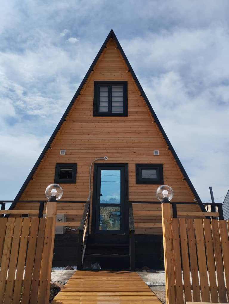The A-frame eco-house is presented with a striking triangular facade, featuring natural wood siding and large windows. A welcoming entrance is framed by a wooden fence and pathway leading to the main door, accentuated by modern light fixtures.