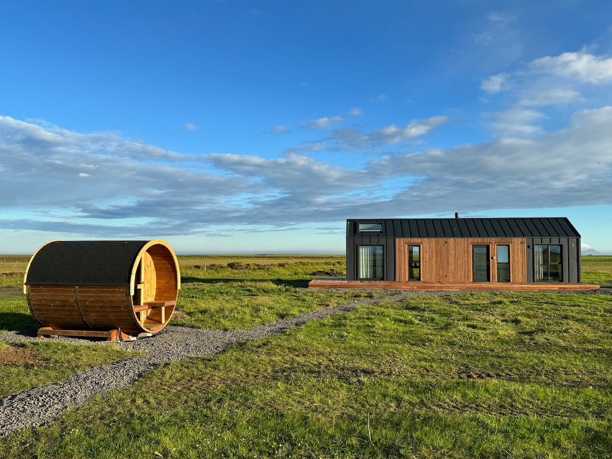 A modern villa is positioned beside a traditional wooden sauna, both surrounded by expansive green fields under a clear blue sky. The villa features large windows providing ample natural light and views of the tranquil landscape.