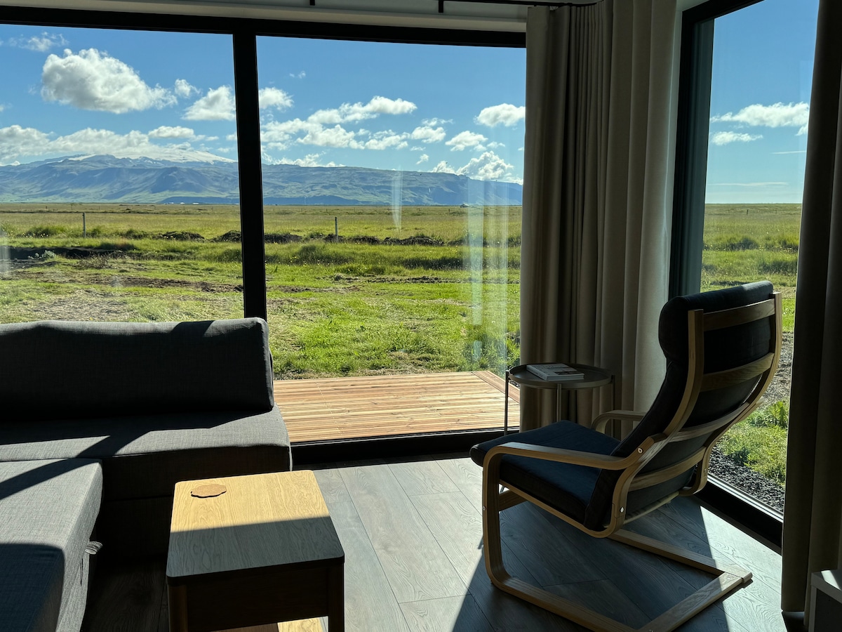 A spacious living area is captured, featuring a modern couch and a wooden side table. Large glass windows offer expansive views of the lush landscape and distant mountains, allowing natural light to fill the room.