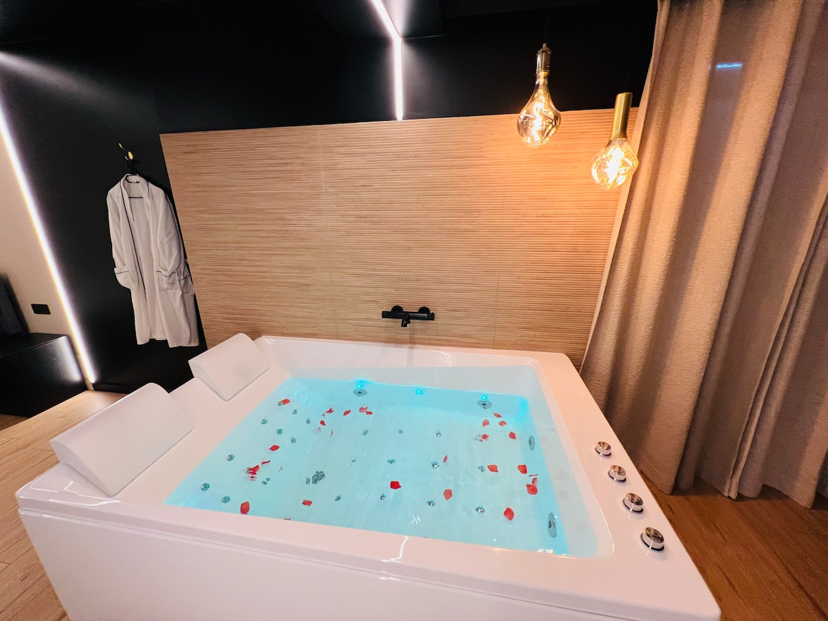 A luxurious spa bath is filled with water and scattered rose petals, creating a soothing ambiance. Soft lighting from pendant lamps reflects off the water's surface, while a plush bathrobe hangs nearby. The elegant design features clean lines and natural wood accents.