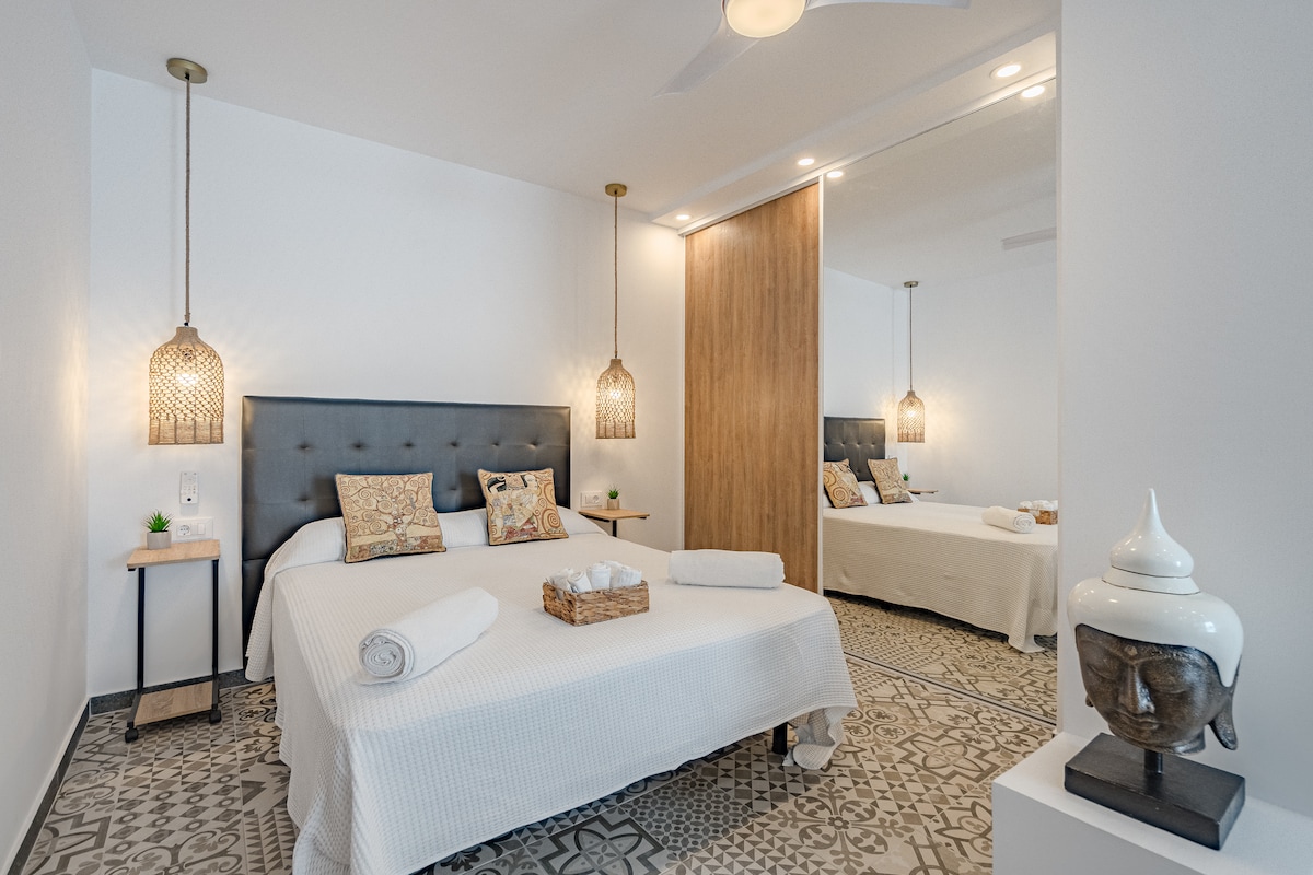 A modern studio bedroom features a sleek bed with decorative pillows and accent lighting from woven pendant lamps. A mirror extends across one wall, enhancing the sense of space. A wooden nightstand with a small plant is positioned beside the bed, complementing the earthy tones of the design.