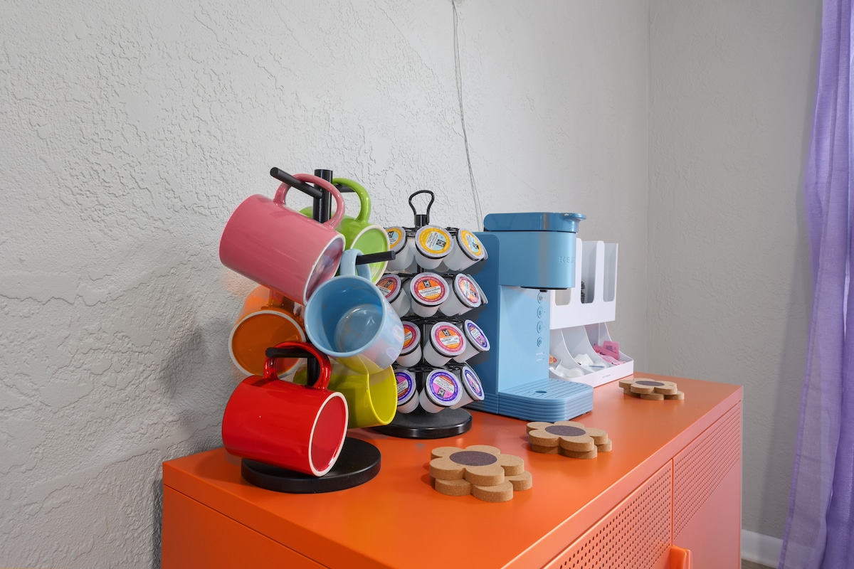 A modern coffee station displays a variety of colorful mugs and a single-serve coffee machine. The mugs are arranged on a tiered holder, while a selection of coffee pods is visible. The orange countertop provides a vibrant contrast to the soft grey walls.