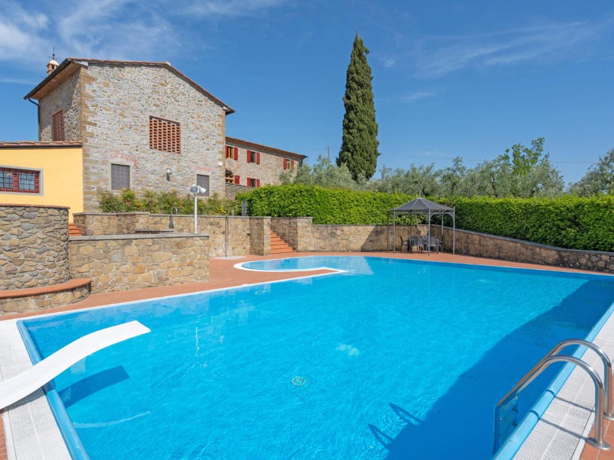 A large, inviting swimming pool is surrounded by lush greenery and stone walls. A diving board extends over the water, while a gazebo is visible in the background, offering shade. The rustic stone buildings and vibrant blue sky enhance the serene setting.