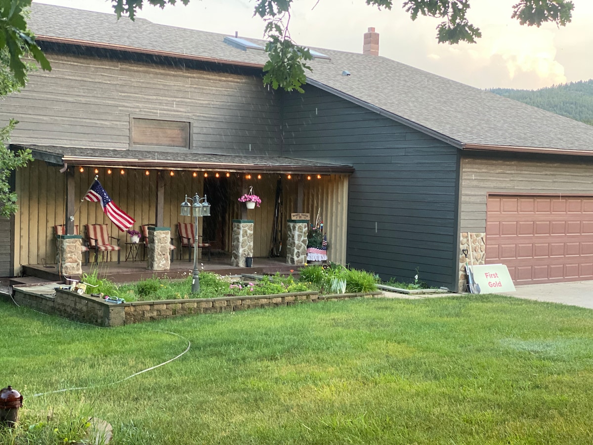 The exterior of the house features a welcoming porch adorned with chairs and decorative elements, including an American flag. Lush green grass surrounds the foundation, and a two-car garage is visible in the background. The home is complemented by natural landscaping and soft outdoor lighting.