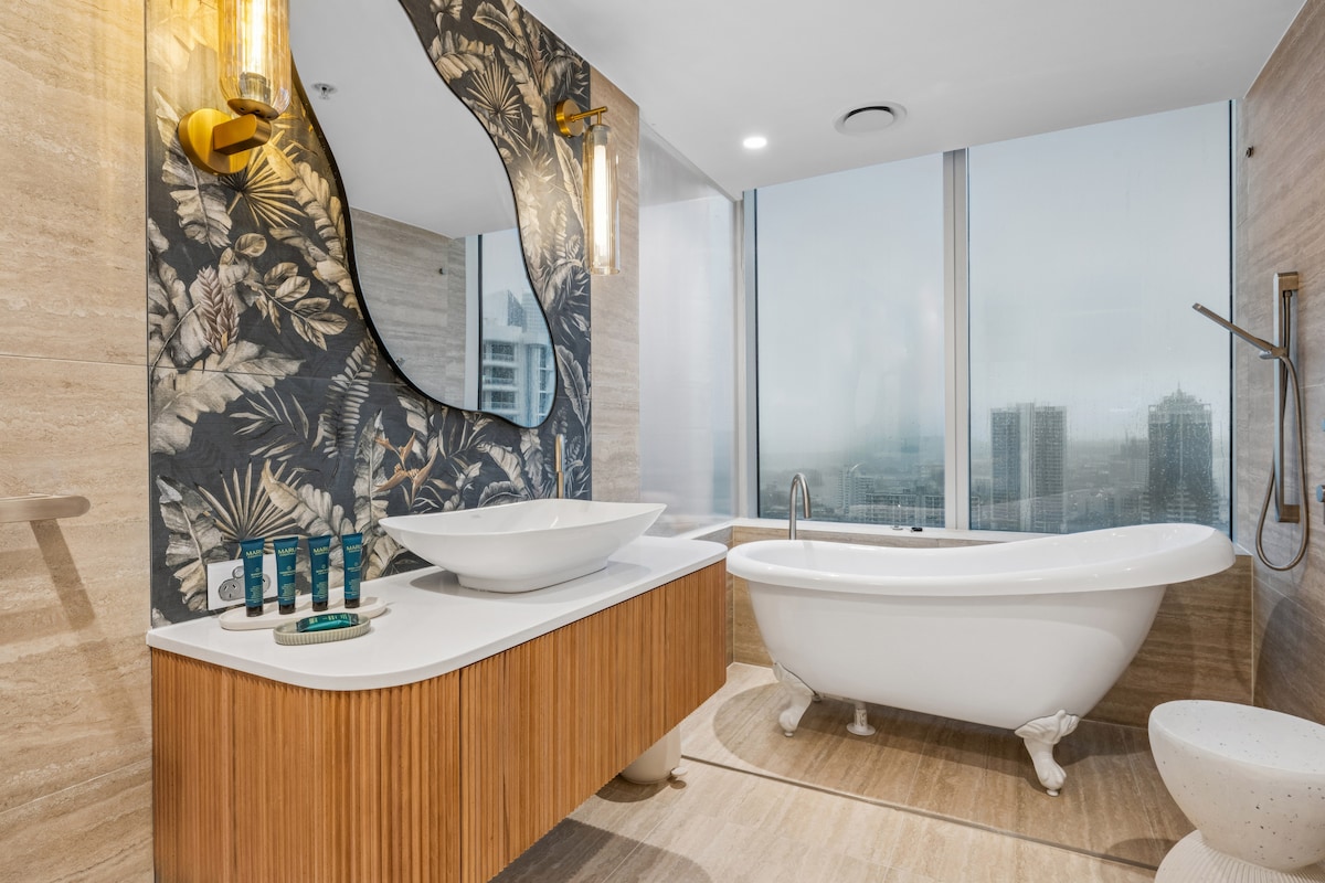 A modern bathroom features a freestanding bathtub and a wall-mounted rain shower. A stylish sink is set on a wooden vanity, while a landscaped wall decor adds a natural touch. Floor-to-ceiling windows provide a view of the city skyline.