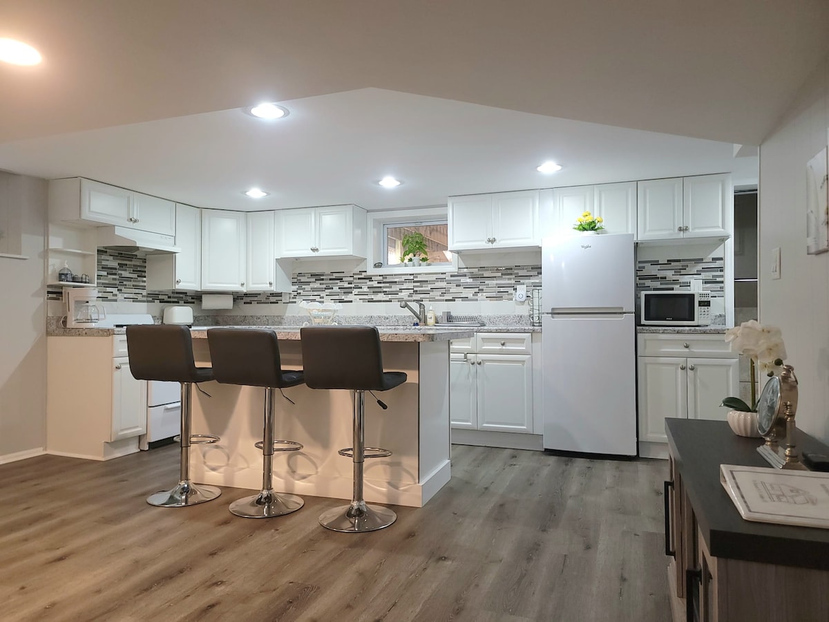 The kitchen is designed with modern white cabinetry and a decorative backsplash. A large island with three barstools provides casual seating. Stainless steel appliances, including a refrigerator and microwave, are featured, while natural light brightens the space from a nearby window.