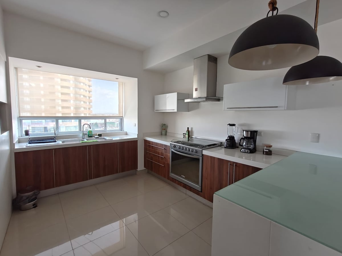 A modern kitchen is showcased, featuring wooden cabinets and white countertops. Stainless steel appliances, including a large oven and coffee maker, are visible. Natural light enters through a large window, highlighting the spacious layout and clean design. Minimal decor contributes to a contemporary aesthetic.