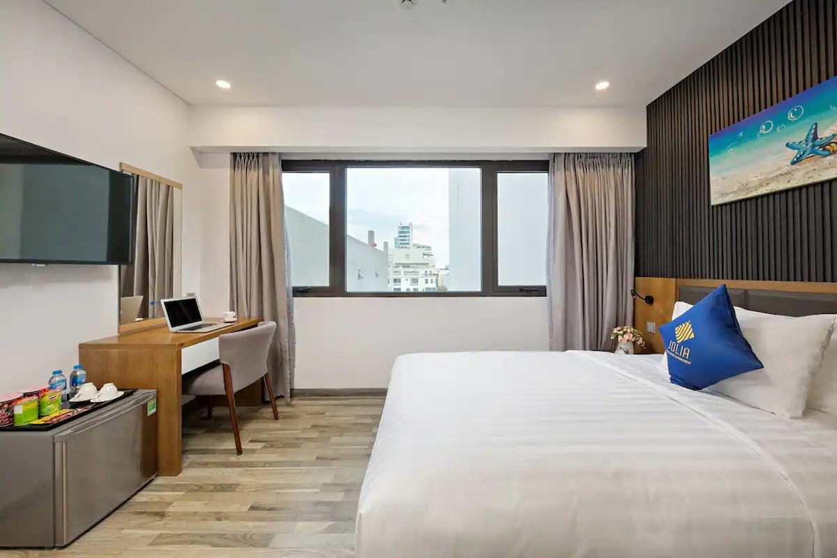 A room featuring a spacious bed with crisp white linens is illuminated by natural light from large windows. A workspace with a desk and chair is present, alongside a mini-fridge. Light-colored wood flooring enhances the modern design, complemented by soft drapes.