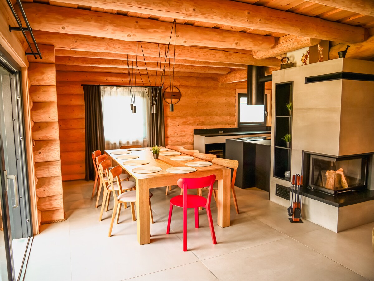 A spacious dining area features a large wooden table surrounded by colorful chairs. Natural light enters through large windows decorated with dark curtains. A modern kitchen is visible in the background, along with a sleek fireplace, adding warmth to the inviting setting.