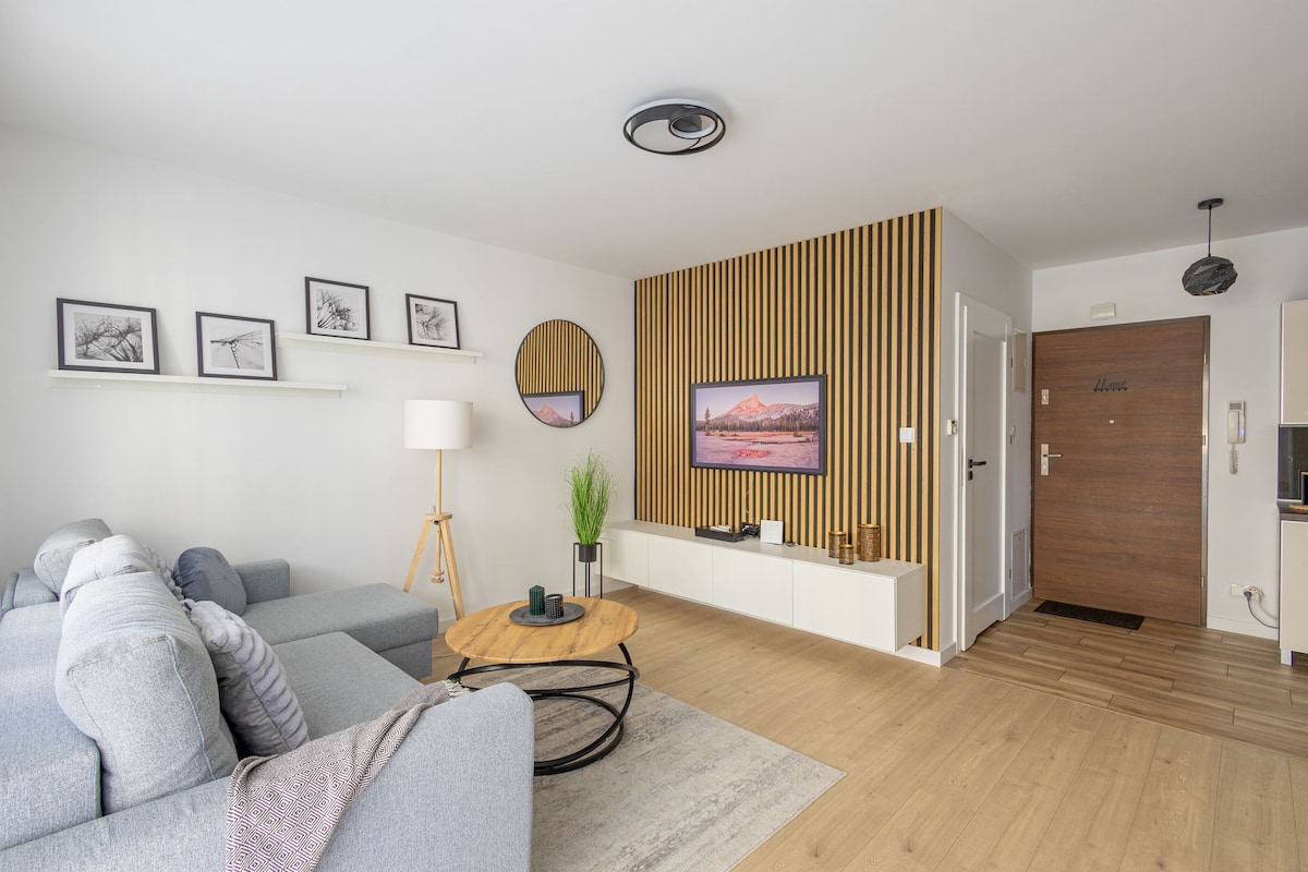 A bright living room features a comfortable sofa and a circular coffee table. Framed artwork adorns the wall, while a round mirror reflects light from the window. A potted plant adds a touch of greenery, and the entrance door is visible in the background.
