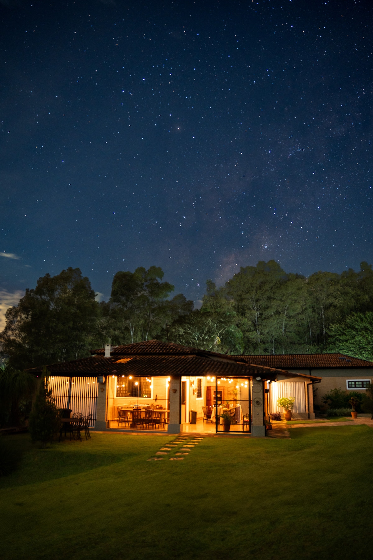 The exterior of the house is illuminated against a starry night sky, showcasing a warm atmosphere. The structure features a spacious porch with seating and a landscaped garden leading to the entrance, framed by tall trees in the background.