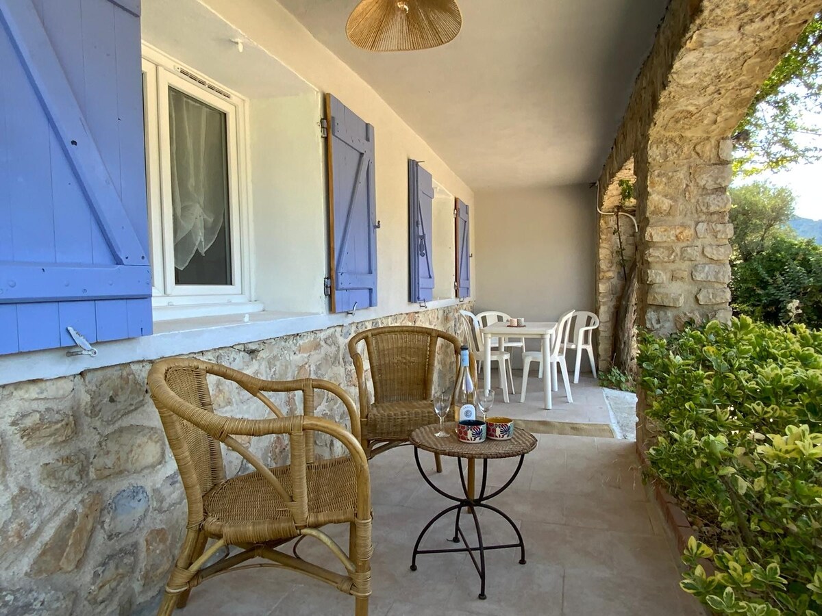 A covered terrace is presented with two woven chairs and a small table. Light blue shutters frame the windows, and stone walls add texture to the space. Lush greenery is visible, enhancing the outdoor setting.