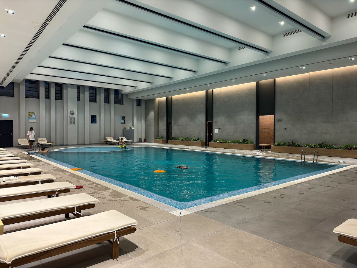 An indoor swimming pool is surrounded by lounge chairs, creating a spacious area for relaxation. The clear water reflects the light from the overhead fixtures, and small flotation devices are visible in the pool. Decorative plants line the edges of the pool area.