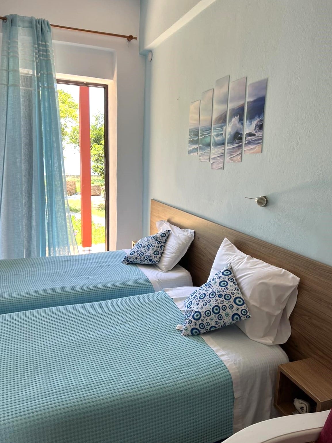 The studio features two single beds with light blue bedspreads, positioned against a light wood panel accent wall. Decorative cushions are placed on each bed. Large windows offer a view of the greenery outside, complemented by sheer blue curtains that gently diffuse natural light.