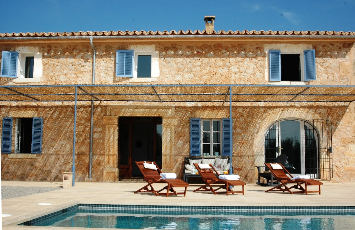 A stone exterior displays traditional Mediterranean architecture, featuring blue shutters and a covered terrace. Four sun loungers are positioned by the pool, offering a space for relaxation. The entrance reveals an inviting interior, blending rustic charm with modern comforts.