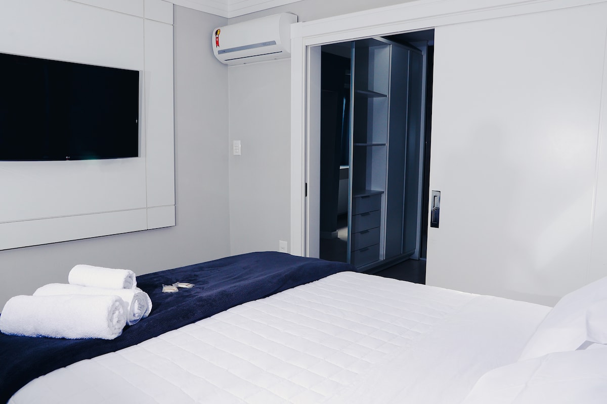 A spacious sleeping area is presented, featuring a neatly made bed with a white quilt and dark blue accent blanket. Soft towels are rolled and placed at the foot of the bed. A wall-mounted TV and open closet space are visible in the background, providing a functional layout.