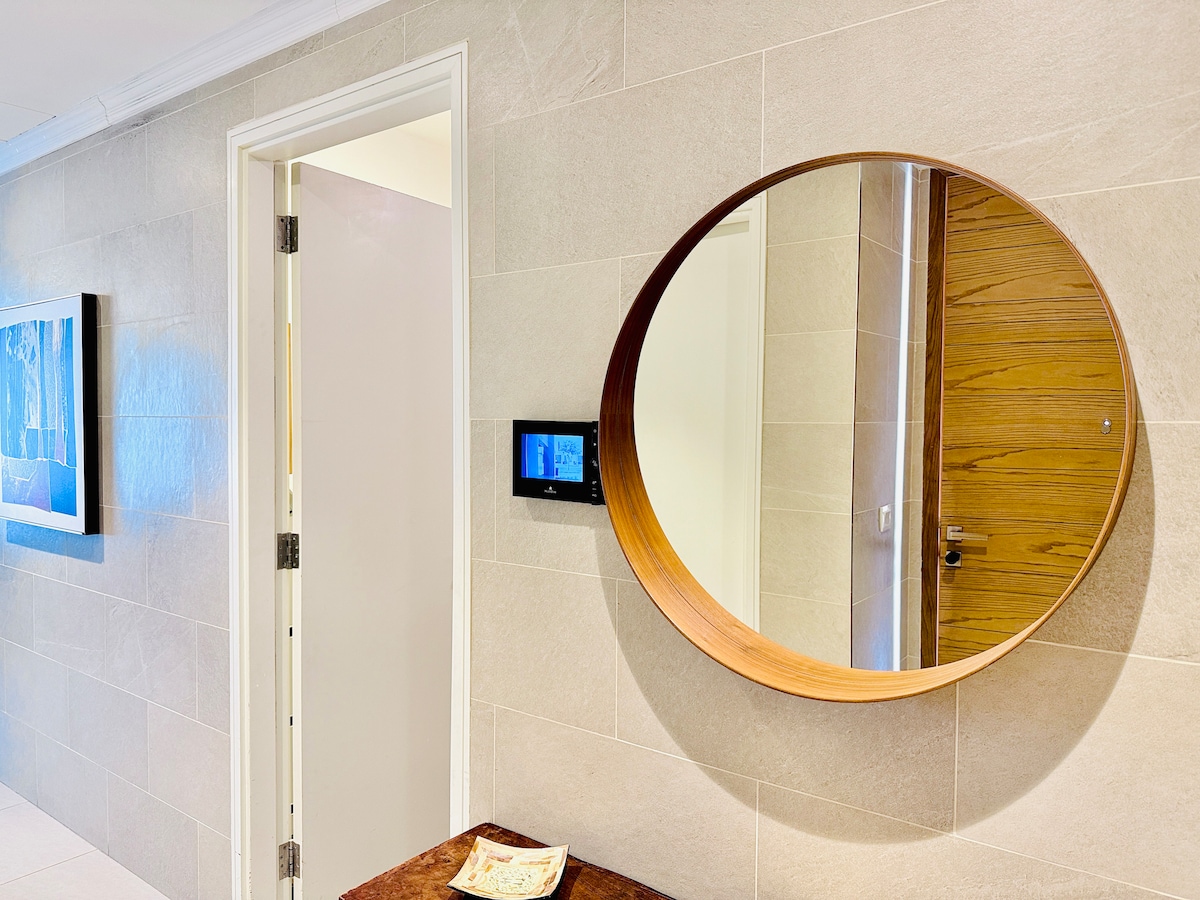 A circular mirror with a wooden frame is mounted on a light-colored tiled wall. A modern digital panel displaying various settings is located beside the mirror. A partially open door is visible in the background, leading to a room with a wooden door.
