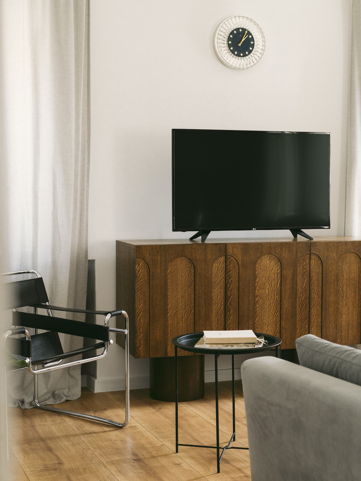 A stylish living area features a sleek TV placed on a wooden cabinet with textured details. A small round table holds a stack of books, accompanied by a modern black chair. Light-colored curtains frame the window, complementing the warm wooden floor.