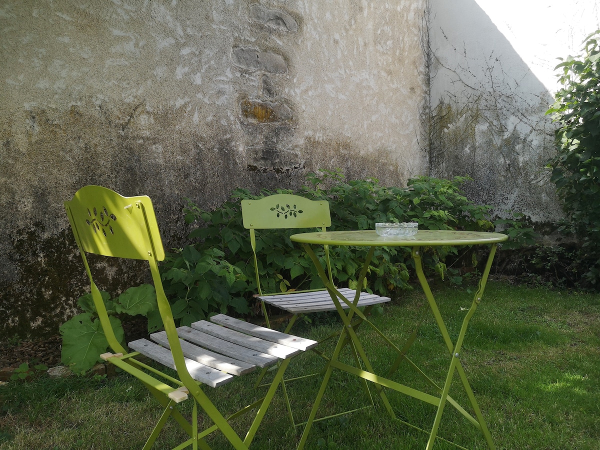A quaint outdoor seating area features a small round table surrounded by two folding chairs. The furniture is set on a grassy patch, with an aged stone wall in the background and greenery providing a natural border.