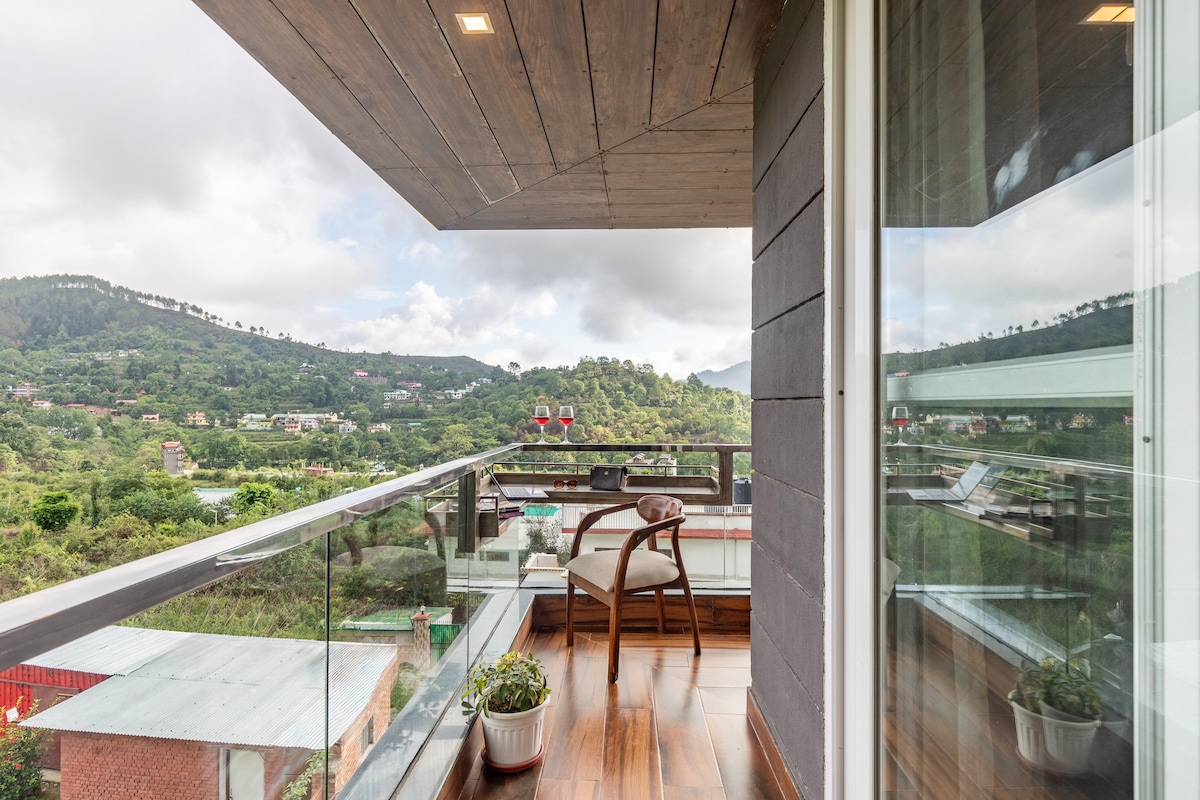 A private balcony overlooking lush greenery offers scenic views of the surrounding hills. A wooden chair along with a small table is positioned against a backdrop of the expansive landscape. Potted plants add a touch of greenery, creating a serene outdoor retreat.