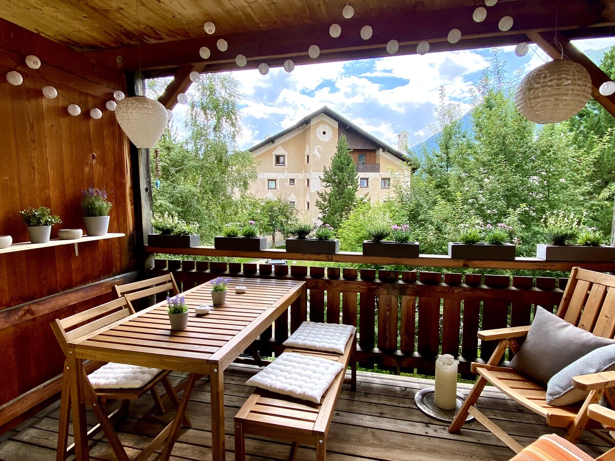 A cozy balcony setting is displayed with wooden furniture, including a table and seating. Flower pots line the railing, and decorative lanterns add a light touch. The surrounding greenery and mountains create a serene atmosphere, with a charming building visible in the background.