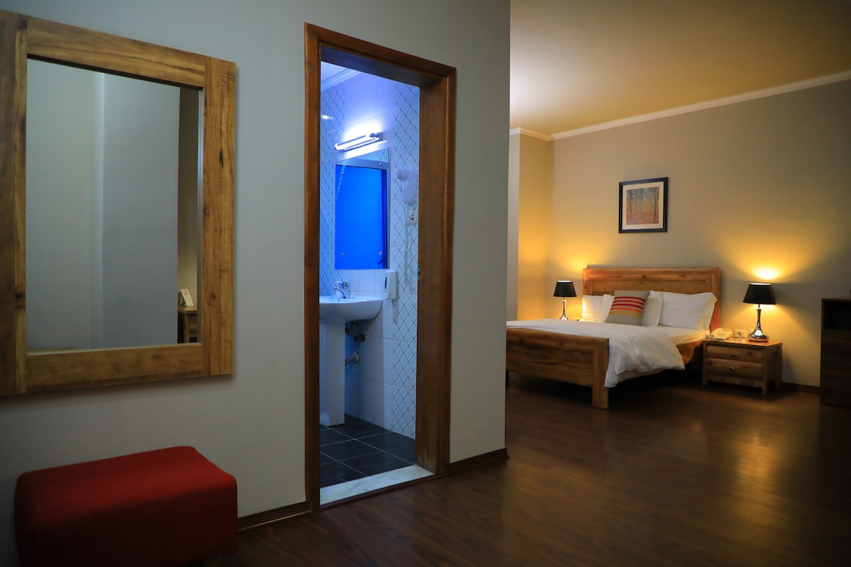 A spacious studio apartment is highlighted, featuring a wooden bed frame with white bedding. A private bathroom doorway is visible, showcasing modern fixtures. A large mirror and a small red ottoman provide additional elements, with bedside lamps softly illuminating the room.