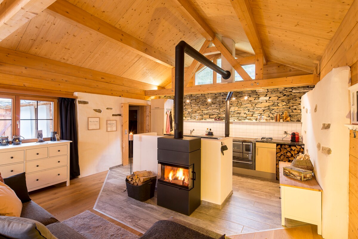 A modern kitchen and living area feature a central wood-burning stove, surrounded by stone accents. The open layout allows natural light to fill the space, with a cozy sofa positioned nearby. Ample cabinets and counter space indicate a well-equipped kitchen for meal preparation.