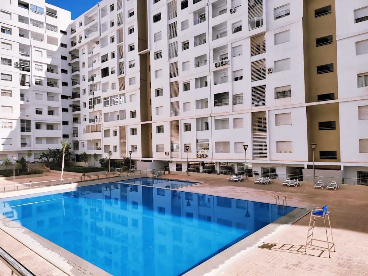 A well-maintained outdoor pool area is featured, surrounded by a spacious deck with lounge chairs. The building's modern architecture is visible around the pool, with numerous windows offering a glimpse of the apartment’s design and natural light.