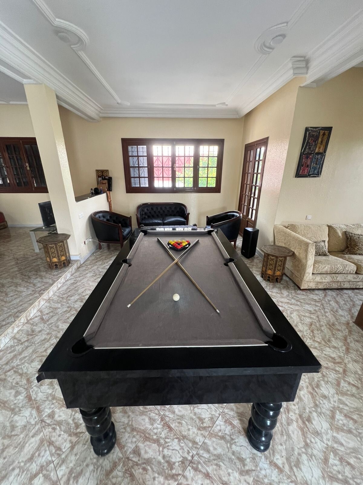 A spacious game room features a sleek black billiard table at its center, accompanied by cues and a set of balls arranged for play. The background includes comfortable seating areas with various chairs and soft armchairs, complemented by wall windows allowing natural light to fill the space.