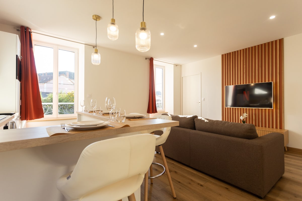 A warm and inviting living space features a spacious sofa oriented towards a wall-mounted television. Modern pendant lights hang above a dining area with elegant table settings. Large windows allow natural light to fill the room, complemented by soft curtains.
