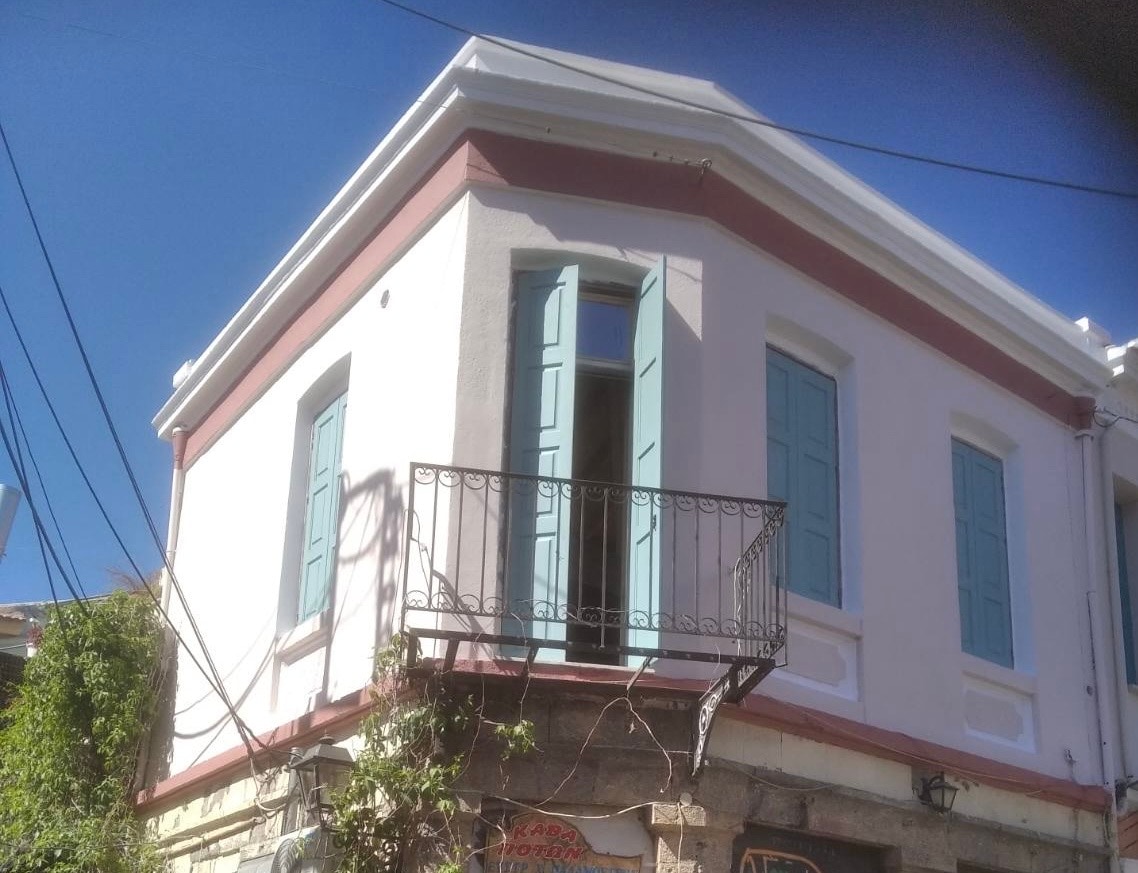 A corner building features a light-colored façade with mint green shutters and a decorative balcony. The structure exhibits a charming architectural style, highlighting its classic design elements and proximity to the vibrant surroundings.