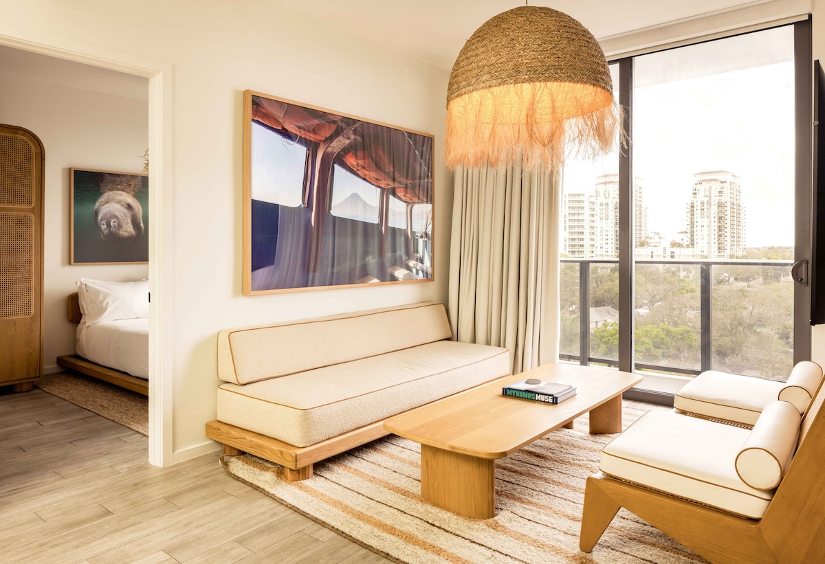 A welcoming living area features a light-colored sofa and matching armchairs, with a wooden coffee table positioned centrally. A large artwork adorns the wall, while natural light flows through the floor-to-ceiling windows leading to a private balcony. A doorway provides glimpses of the adjacent bedroom.