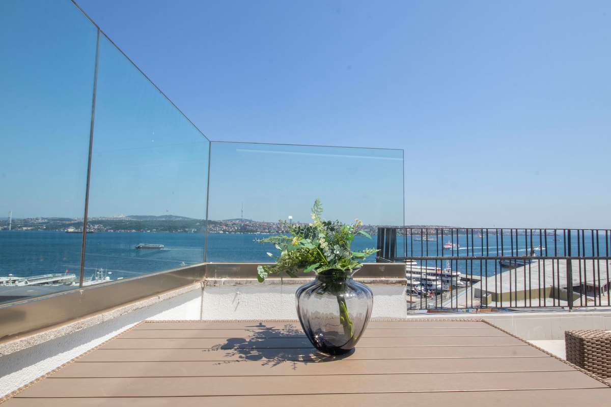 A terrace is presented with a clear view of the water, featuring a table topped with a glass vase containing fresh greenery. The surrounding railing is made of glass, allowing for unobstructed sightlines to the serene landscape beyond.