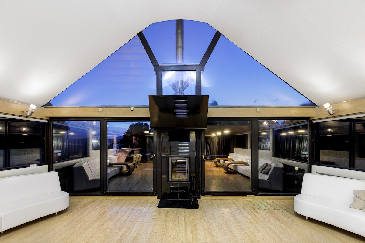 An expansive living area features large glass windows forming a triangular shape, allowing natural light to fill the space. Two modern white sofas are positioned facing a central fireplace, with a mounted TV above. The serene atmosphere is complemented by a view of the evening sky.