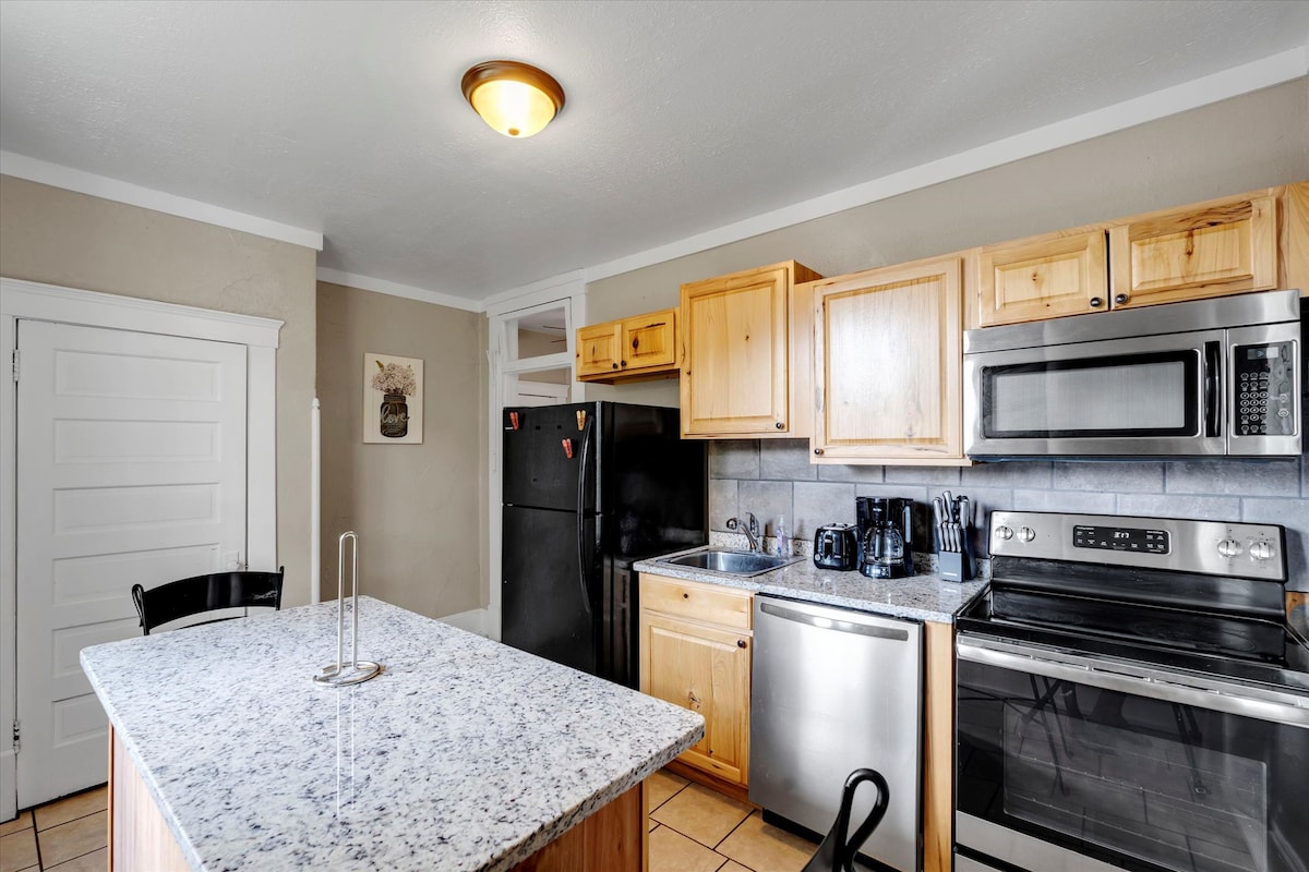 The kitchen features light wood cabinetry and a spacious granite countertop island. Stainless steel appliances are present, including a refrigerator, dishwasher, and stove. Natural light is provided by a nearby window, illuminating the tiled floor and the minimalist decor.