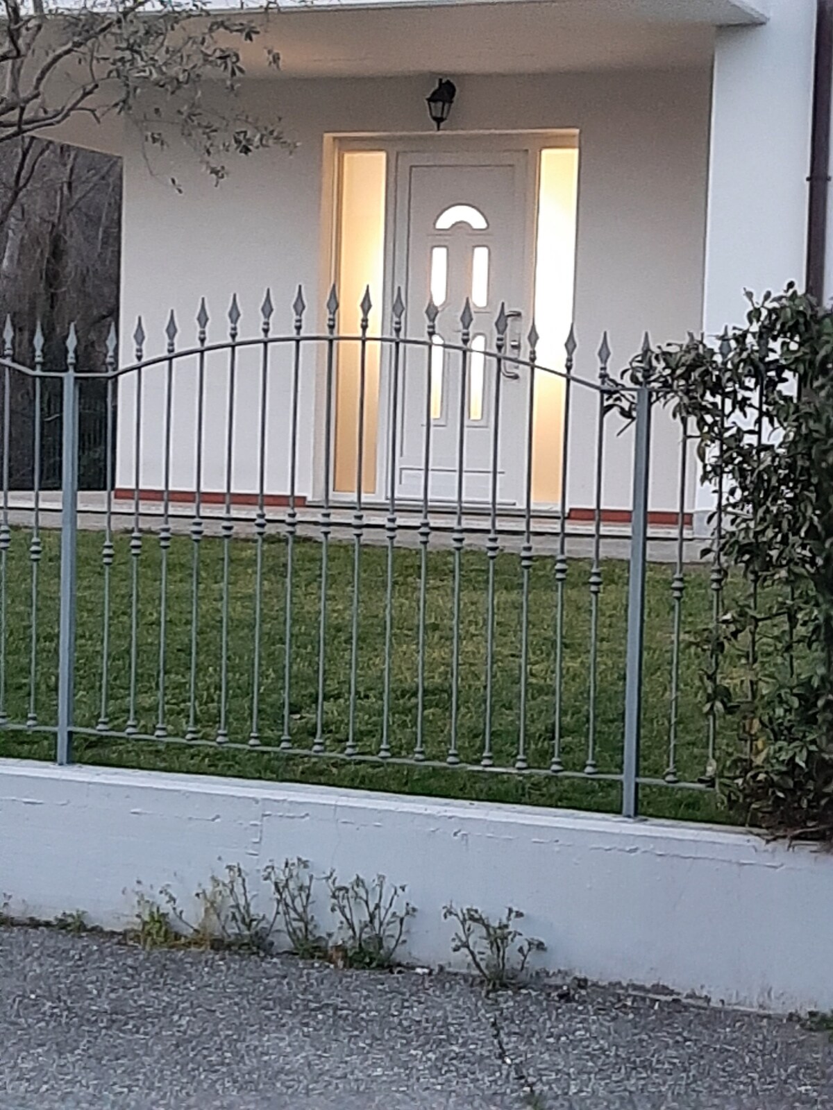 A modern entrance is framed by a decorative iron fence. The well-maintained lawn in front leads to a white door with distinctive window cutouts, softly illuminated from within, providing a welcoming appearance.