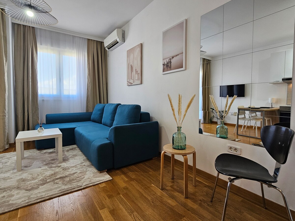 A modern living area features a blue sectional sofa, a small white coffee table, and a minimalist chair. Light-colored curtains frame a large window, providing ample natural lighting. A decorative vase with dried plants adds a touch of warmth, and a mirror reflects the inviting space.