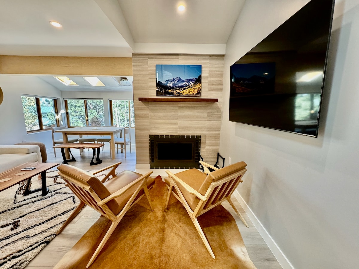 Two wooden chairs with angled backs are positioned in front of a sleek fireplace, accompanied by a soft area rug. A modern TV is mounted on the wall above the fireplace, while a large window with surrounding greenery allows natural light to fill the space.
