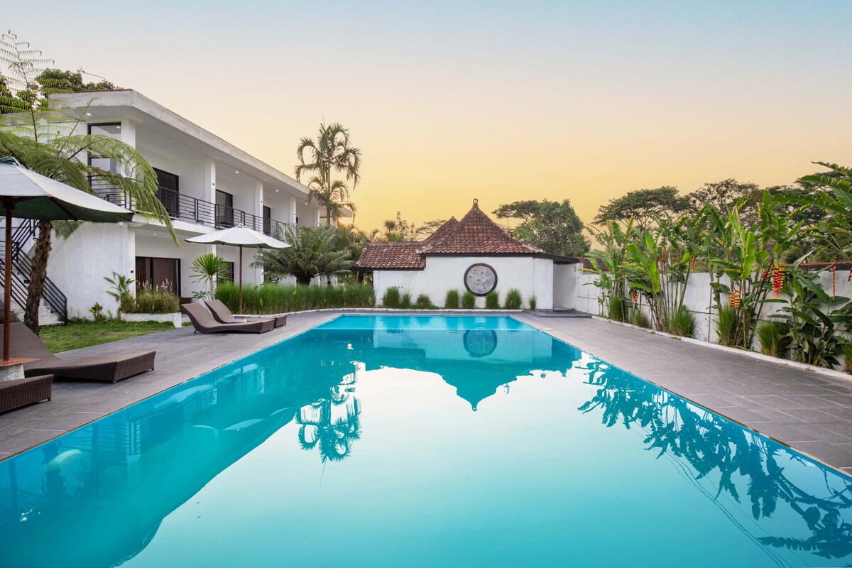 The outdoor area features a sparkling swimming pool, reflecting the sky in tranquil blue tones. Comfortable lounge chairs are positioned beside the pool, surrounded by lush greenery and tropical plants. A traditional structure can be seen in the background, enhancing the serene setting.