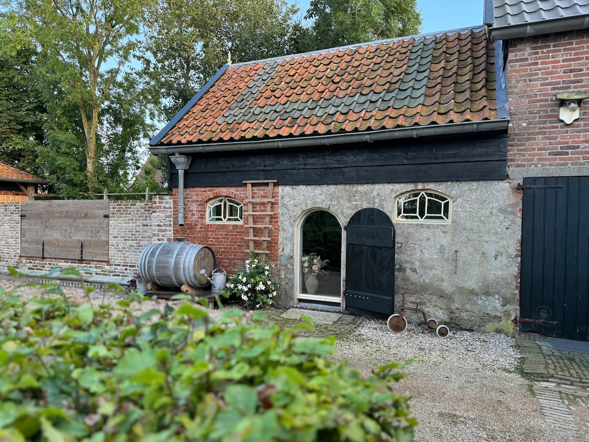 A rustic facade of a barn apartment is presented, featuring a combination of stone and wood materials. Arched windows provide an inviting entrance, framed by greenery. A decorative barrel and gardening elements enhance the exterior, set against a backdrop of trees and a gravel pathway.
