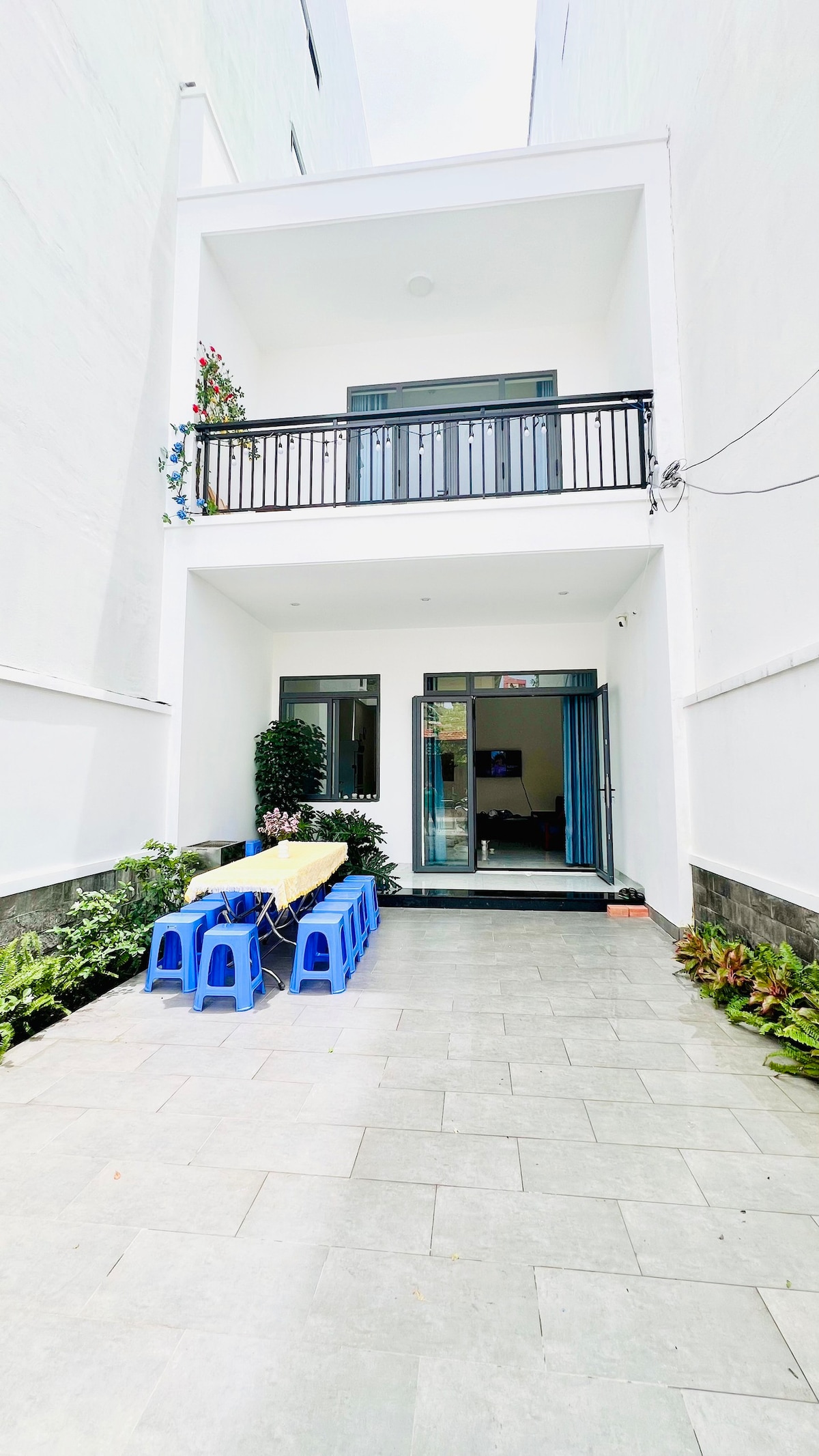 An outdoor area is featured, featuring a long table set with blue chairs. The space is bordered by greenery and is illuminated by natural light. A balcony above overlooks the area, with flowers cascading down, enhancing the inviting atmosphere.