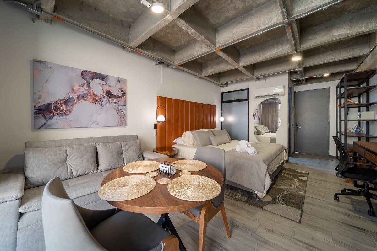 A modern spacious loft features a queen bed and a double sofa bed. A round dining table with four chairs sits on a patterned rug. Natural light enters through a large window, enhancing the room's neutral color palette and industrial ceiling design.